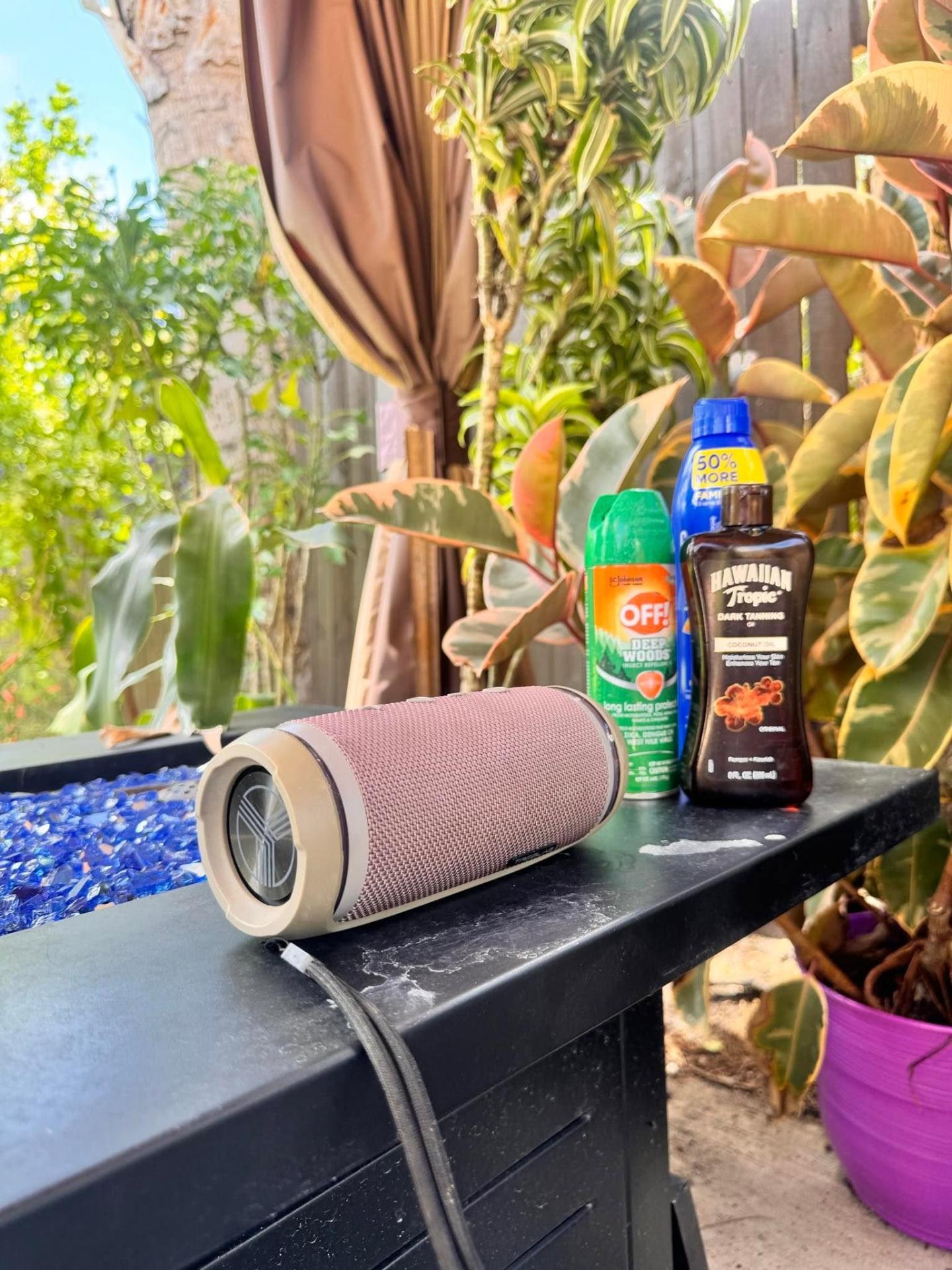Bluetooth speaker, sunscreen and tanning oil on a poolside table beside a fire feature